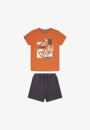 Camiseta naranja de manga corta con gráfico fragmentado blanco de león y texto "Jungle in Wild", combinada con pantalones cortos gris oscuro con cordón.