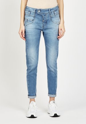 Femme portant un jean skinny bleu clair retroussé et des chaussures de sport blanches, debout devant un fond uni.