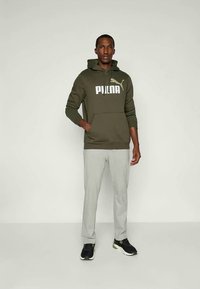 Felpa con cappuccio verde oliva con logo "PUMA" bianco, abbinata a pantaloni grigi chiari a fantasia e scarpe da ginnastica nere con dettagli verdi.