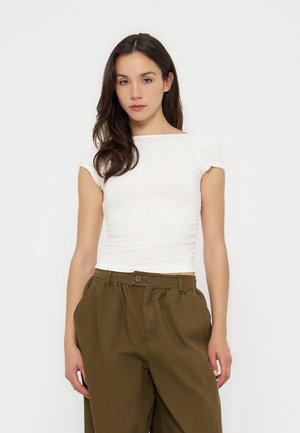 Femme aux longs cheveux foncés portant un haut blanc ajusté à manches courtes et un pantalon ample vert olive, debout devant un fond uni.