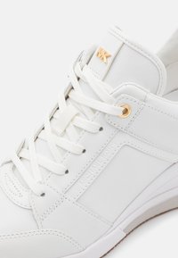 Hvit sneakers med en glatt lær- og tekstiloverdel, flate lisser, gull aksent og preget logo-detalj på siden.