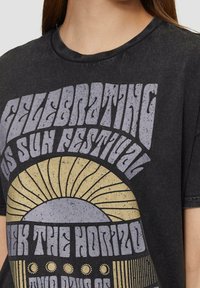 Osoba oblečená v černém vintage tričku s grafikou slunce a textem "Oslava festivalu Los Sun" a "Projděte horizontem."