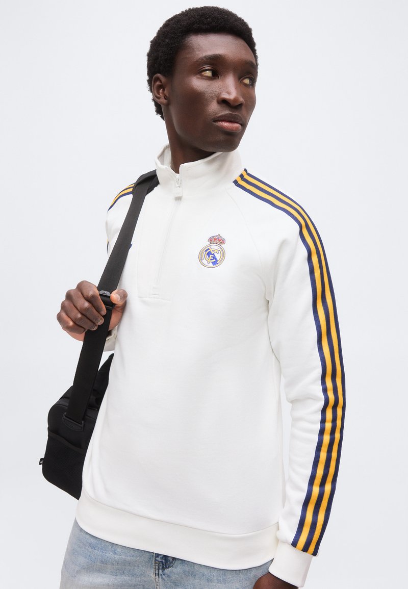 Jeune homme portant un pull blanc Real Madrid à fermeture éclair jusqu'au quart avec des bandes bleues et jaunes sur les manches, tenant la sangle d'un sac à bandoulière noir.