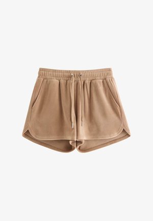 Ung kvinde iført en beige kasket, briller, hvid strikket cardigan, hvid top, brune shorts, hvide sokker og hvide sneakers, stående mod hvid baggrund.