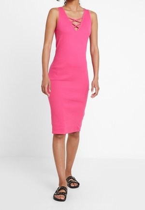 Femme portant une robe sans manches rose vif longueur genou avec un détail croisé au niveau du décolleté et des sandales noires cloutées.