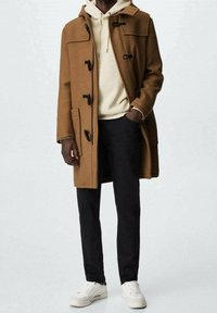Cappotto di lana marrone con cappuccio, dotato di chiusure a toggles, due tasche frontali, indossato sopra un hoodie crema, abbinato a pantaloni neri e sneakers bianche.