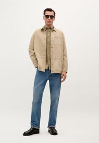 Giacca di cotone beige con tasche frontali sopra una camicia verde chiaro con bottoni, abbinata a jeans blu e mocassini neri, indossata da un modello maschile.