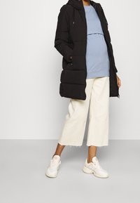 Schwangere Person in einer schwarzen Steppjacke, einem blauen Oberteil, hellbeigen weiten Hosen und weißen Sneakers, die vor einem einfarbigen Hintergrund steht.