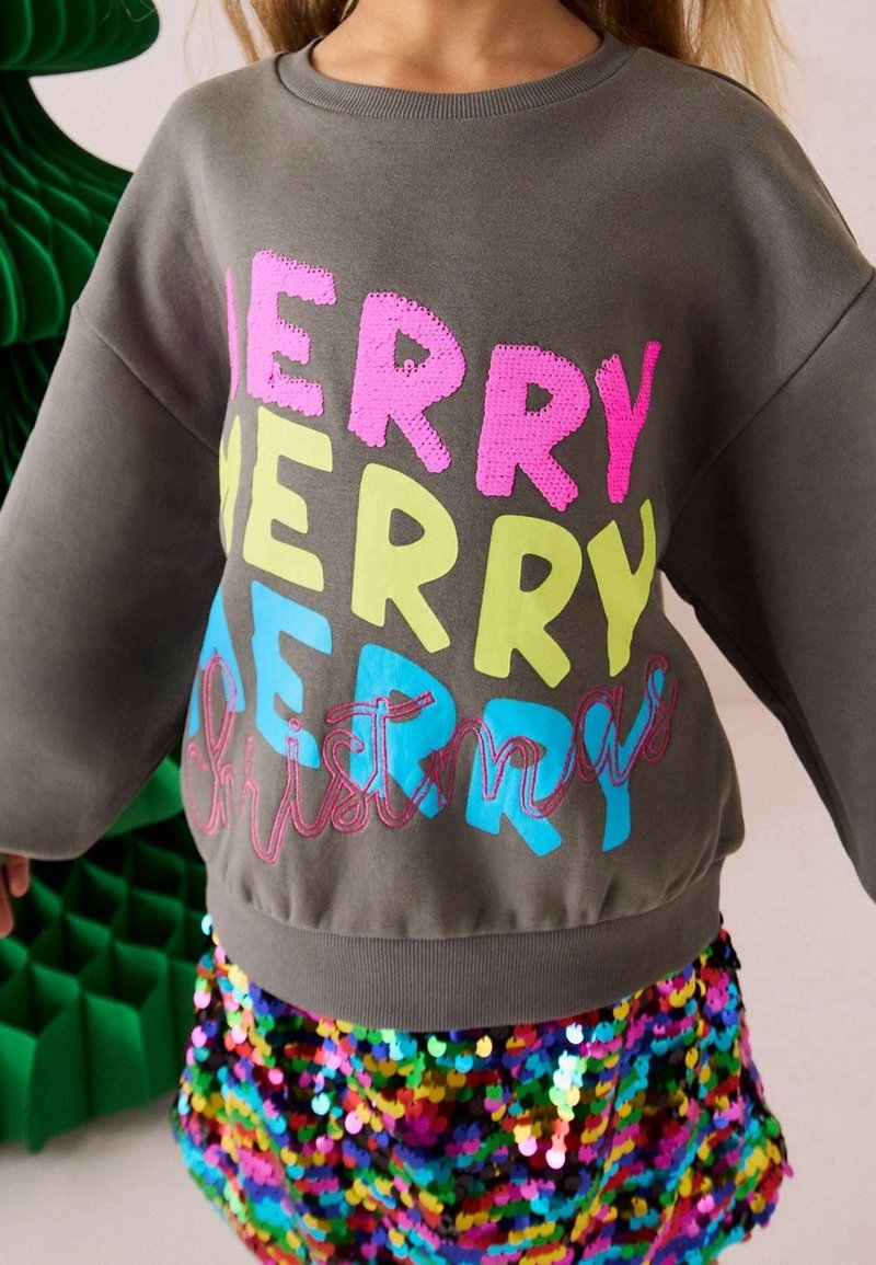 Enfant portant un sweat gris avec le texte coloré "Joyeux Noël" et une jupe pailletée multicolore, debout près d'une décoration verte.