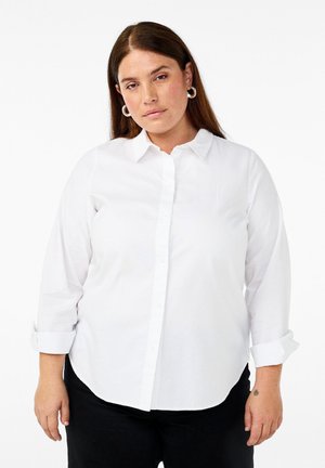 Witte overhemd met lange mouwen, een kraag en opgerolde mouwen, gemaakt van een gladde stof; gedragen met zwarte broek.
