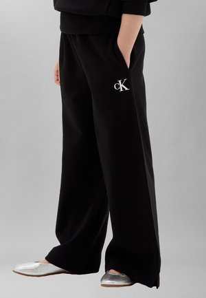 Persona che indossa pantaloni neri a gamba larga con logo bianco "cK" sulla coscia, mani in tasca e scarpe piatte argento, in piedi su sfondo grigio.
