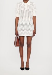 Robe polo blanche en tricot avec des manches courtes et un motif à trous ouverts, accompagnée d'un sac à main marron et de ballerines noires à bout pointu.