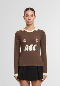 Ruskohvi pitkähihainen paita vaalealla kauluksella ja näkyvällä valkoisella logolla. Yhdistelee mustaan laskostettuun hameeseen. Sileä kangas.