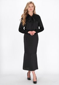 Zwarte satijnen jurk met een nauwsluitend lijfje, lange mouwen en een strik bij de kraag, met een uitlopende rok die tot de enkels valt.