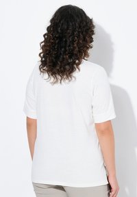 Chemise blanche à manches courtes avec une coupe décontractée. Texture douce, poignets roulés et fentes sur les côtés. Vue de dos, cheveux partiellement visibles.