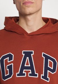 Sudadera color burdeos con letras "GAP" en blanco y azul marino. Cuenta con capucha ajustable y una textura suave. El corte es casual y desestructurado.