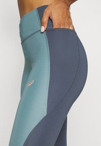 Høyt midje leggings med to-farget design i teal og marineblå, glatt tekstur, kontrasterende sømmer, og en liten merkevarelogo på låret.