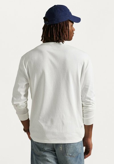 Camicia bianca a maniche lunghe con scollatura rotonda e polsini a coste, abbinata a jeans di denim. Il modello indossa un cappellino blu navy, rivolto lontano dalla telecamera.