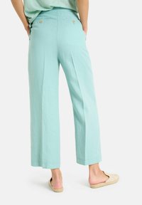 Licht turquoise wijde broek van zacht materiaal, met een hoge taille en twee knopen aan de achterkant. Het model draagt beige instapschoenen.