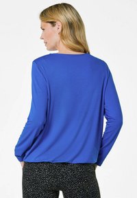 Saphirblaues Langarmoberteil mit lockerer Passform, rundem Ausschnitt und glatter Textur, kombiniert mit schwarzen gemusterten Hosen.