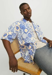Korte mouwen shirt met een blauw en crème hexagonaal patroon, gemaakt van lichte stof, gedragen over een beige shirt, gecombineerd met spijkerbroeken.
