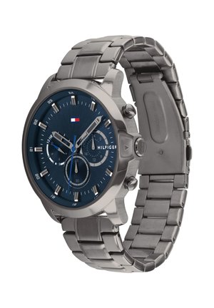 Reloj de pulsera de metal plateado con esfera azul, tres subesferas, logo de Tommy Hilfiger y pulsera enlazada con cierre de broche.