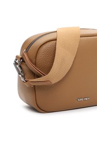Bolso bandolera de cuero marrón texturizado con correa ancha de tela beige y cremallera plateada, mostrando el pequeño logotipo de la marca "SURI FREY."