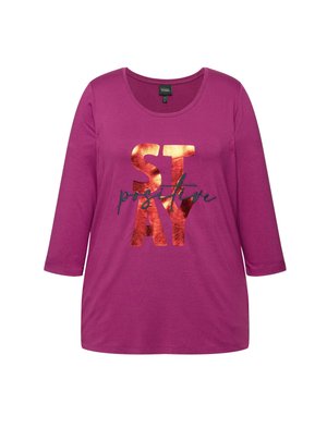 Camiseta magenta de manga larga con escote redondeado y letras negras que dicen "MANTENTE positivo", con acentos metálicos en color cobre en las letras.