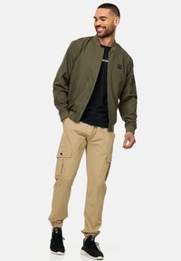 Chaqueta bomber verde oliva con un parche negro, combinada con pantalones cargo beige que tienen bolsillos laterales y puños elásticos, llevada con zapatillas negras.