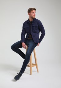 Giubbotto in suede blu con due tasche sul petto, abbinato a jeans scuri e scarpe blu, seduto su uno sgabello di legno contro uno sfondo neutro.