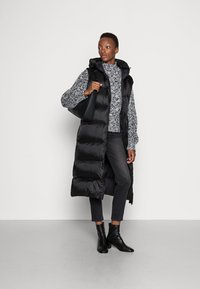 Gilet imbottito nero con cappuccio, caratterizzato da un design trapuntato, indossato sopra un maglione grigio a fantasia, abbinato a jeans scuri e stivaletti neri.