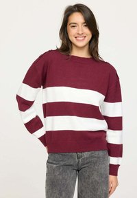 Pull rayé bordeaux et blanc avec un col rond, texture tricotée et accents de boutons aux épaules. Assocé à un jean gris ajusté.