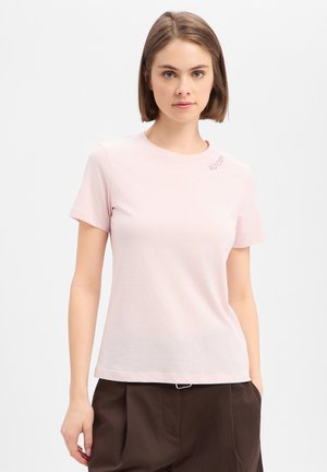 Junge Frau trägt ein hellrosa kurzärmeliges T-Shirt und dunkelbraune Hose, steht mit einer Hand in der Tasche vor einem einfarbigen Hintergrund.