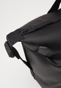 Čierna ramenná taška vyrobená z hladkého, textúrovaného materiálu. Obsahuje zips, nastaviteľný popruh a minimalistický dizajn s jemným brandingom.