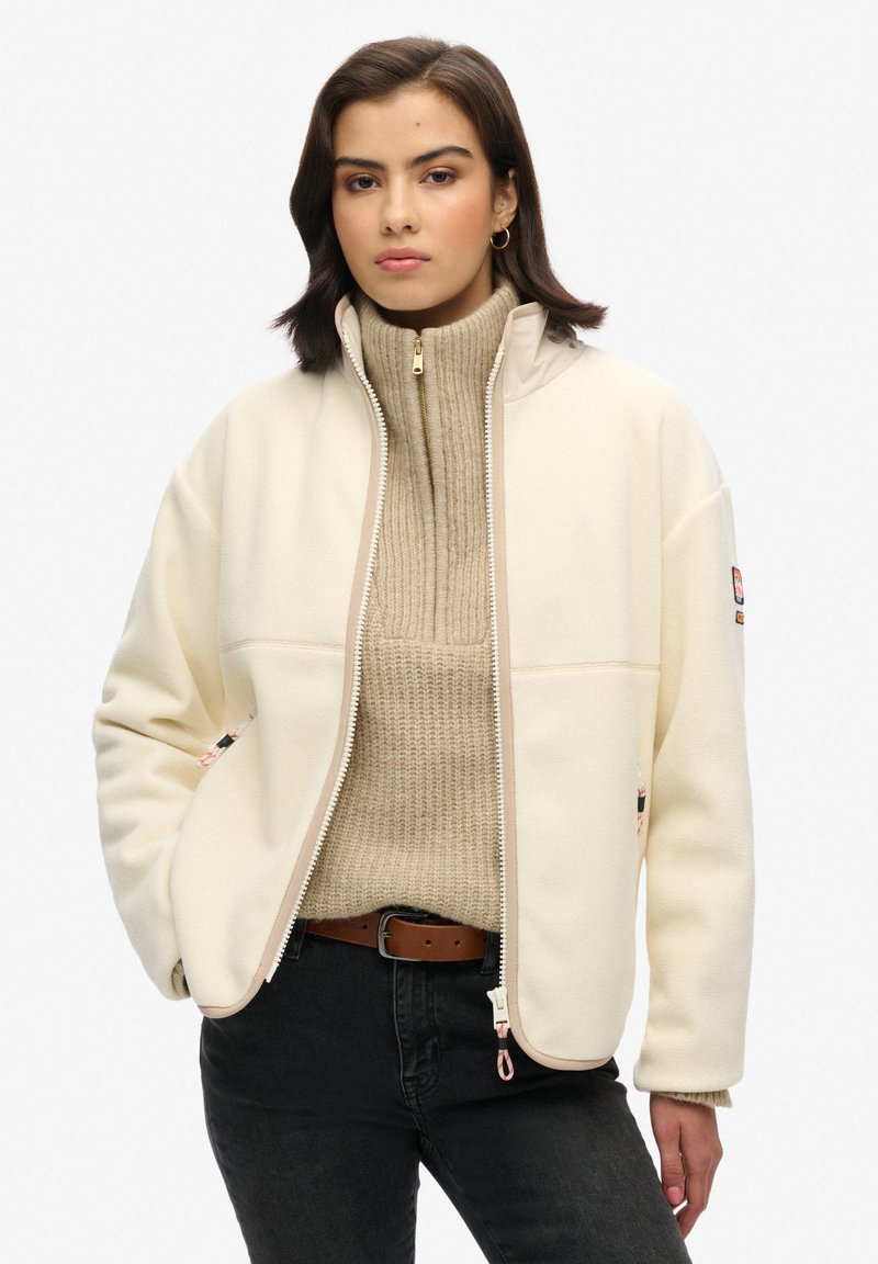 Fleecejack in crèmekleur met een rits aan de voorkant, voorzien van ribben in beige bij de nek en accenten. Inclusief twee zijzakken en een label op de linker mouw.