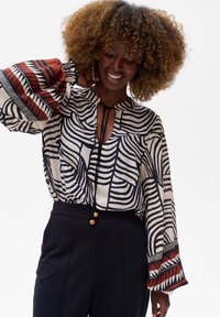 Biała bluzka z czarnymi falistymi liniami, z dekoltem w kształcie łezki, elastycznymi dzwonkowymi rękawami z czerwonymi i czarnymi akcentami geometrycznymi.