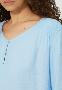 Lichtblauwe blouse met een V-hals, voorzien van een gestructureerde stof, fronsdetails bij de schouders en een enkele knoopsluiting.