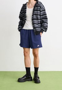 Černá a šedá pruhovaná mikina, bílý tílko, navy šortky, černé platformové boty a černé ponožky, stojící proti neutrálnímu pozadí.