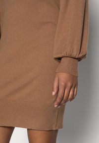 Braun gestricktes Kleid mit gerippten Bündchen und Saum, lässiger Schnitt und strukturierter Oberfläche. Eine Hand ist sichtbar, die glatte Haut und Nägel zeigt.