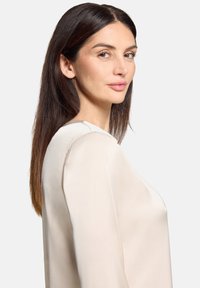 Beige Seidenbluse mit rundem Ausschnitt und langen Ärmeln, zeichnet sich durch eine glatte Textur und einen dezenten Glanz aus. Das Modell posiert in Profilansicht.