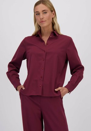 Blouse bordeaux avec un col, une fermeture à boutons sur le devant et des manches longues. Le tissu semble lisse avec une coupe légèrement décontractée.