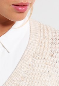 Beige neulepaita, jossa on teksturoidut, avoimet kudontayksityiskohdat ja ribbireunus, valkoisen kauluspaitan päällä. Lähikuvassa korostuu kankaan yksityiskohdat.