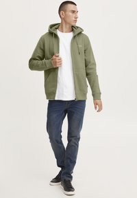 Felpa con zip verde oliva con tasca a marsupio, cappuccio con cordino e polsini a costine, indossata sopra una maglietta bianca e abbinata a jeans blu.