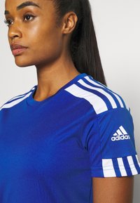 Camiseta deportiva azul con franjas blancas en los hombros y logo de Adidas en la manga. La tela parece suave y ajustada, con mangas cortas y cuello redondo.