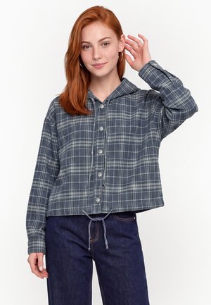Jeune femme aux cheveux roux portant un sweat à capuche gris à carreaux boutonné et un jean bleu foncé, souriant et touchant ses cheveux d'une main.
