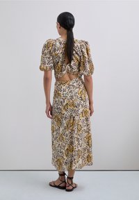 Woman with long tied-back hair wearing a yellow and black patterned open-back top and matching midi skirt with black lace-up sandals.