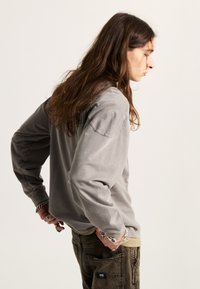 Šedá dlouhá tričko s volným střihem, s kulatým výstřihem a dekorativním prošíváním, spárované s hnědými texturovanými kalhotami.