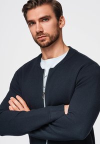 Marinefarbener Zip-Pullover mit geripptem Kragen und Bündchen. Der Stoff wirkt weich und strukturiert. Das Modell hat die Arme gekreuzt und zeigt die Designdetails.