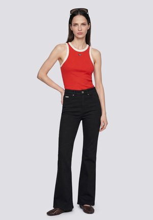 Femme portant un haut rouge sans manches avec une bordure blanche, pantalon noir évasé taille haute, lunettes de soleil sur la tête et ballerines tressées foncées, posant avec une main sur la hanche.