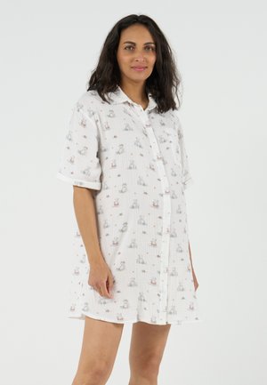 Zwangere vrouw staand, gekleed in een losse witte jurk met knopen en kleine dierenprints, tegen een effen lichte achtergrond.